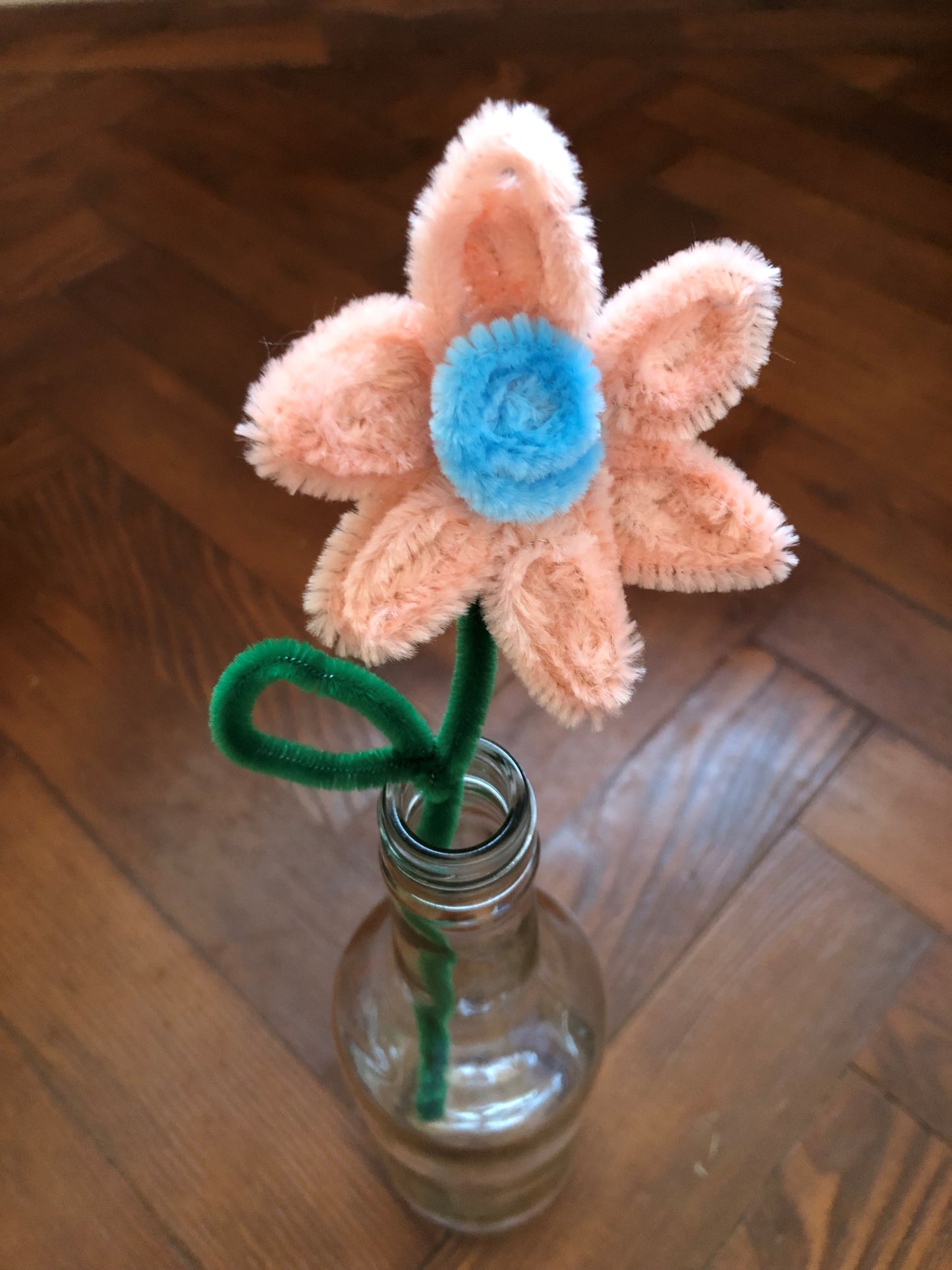 A flower made out of pipe-cleaners sits in a clear glass bottle on a wooden background. The flower is blue and pink, with a green stalk.