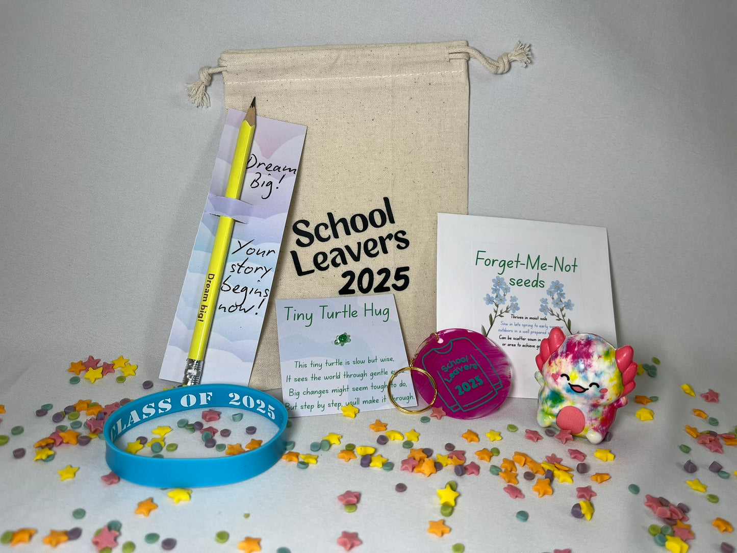 A white canvas bag reading "School Leavers 2025" sits on a confetti background. The contents are shown: a yellow pencil printed with "dream big", a blue wristband that says Class of 2025, a turtle sits on a card with a poem titled Tiny Turtle Hug, a pink circular keyring has a school leavers 2025 hoody on it, there is a packet of Forget-Me-Not seeds, and a rainbow coloured axolotl squishy toy.