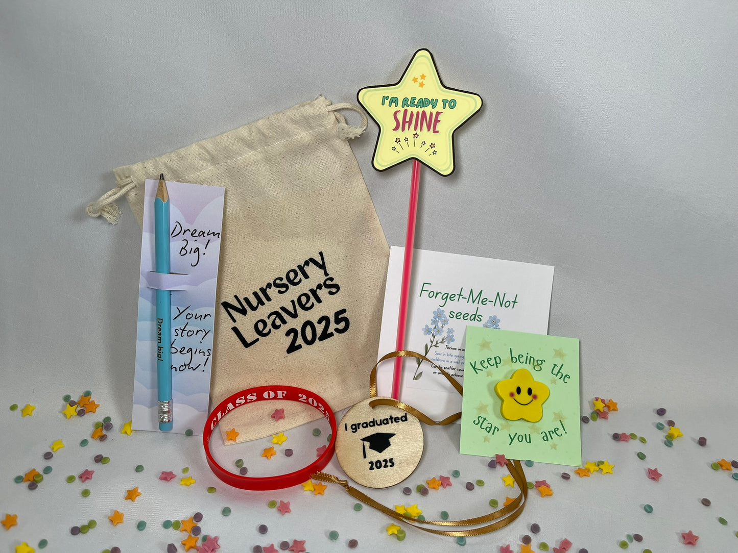 A white canvas bag reading "Nursery Leavers 2025" sits on a confetti background. The contents are shown: a blue pencil printed with "dream big", a red wristband that says Class of 2025, a yellow star rubber with a smile sits on a card titled “keep being the star you are”, a wooden circular medal has a graduation hat on it and the words “I graduated 2025”, there is a packet of Forget-Me-Not seeds. There’s a red glowstick with a star at the top, it looks like a magic wand, and reads Ready to Shine
