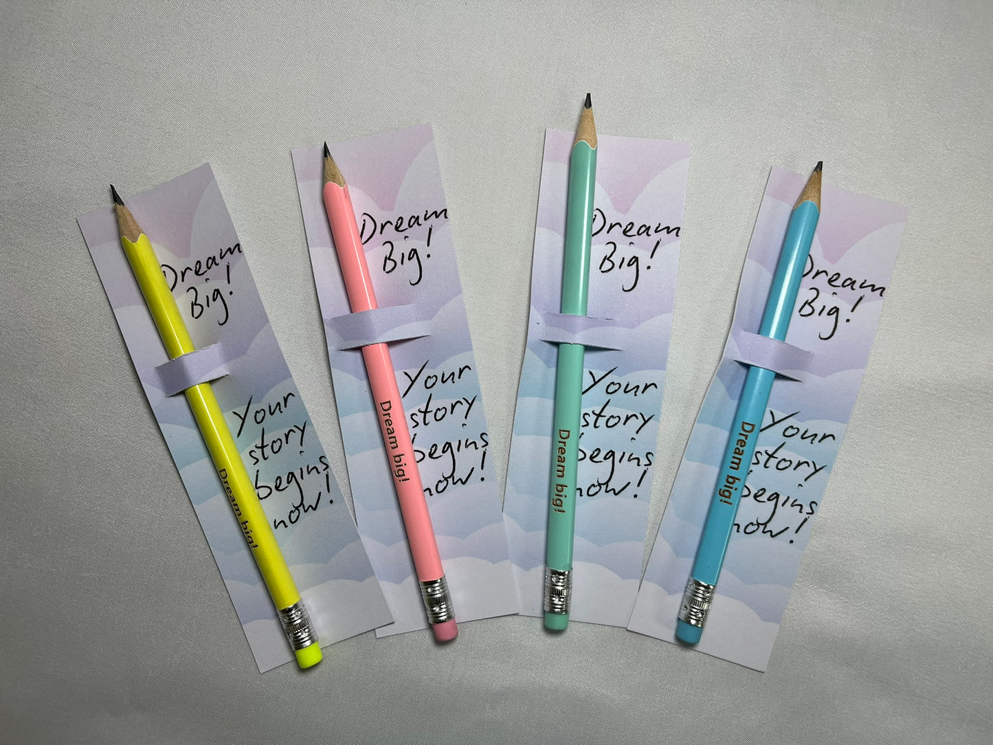 Four pencils are fanned out. They are yellow, pink, pale green and blue. Each pencil slots into a card that reads “Dream Big” Your story begins now!”. The pencils are engraved with the words “Dream Big!”