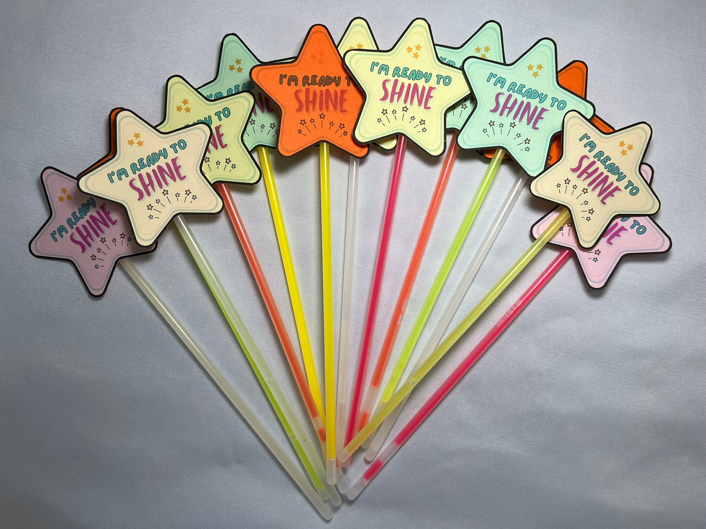 Lots of wands are spread out. Each wand is a glowstick with a star at the top, the star says “I’m ready to shine”. The stars are brightly coloured, pink, yellow, green, peach, orange. The glowsticks are also mixed colours, each wand is different.