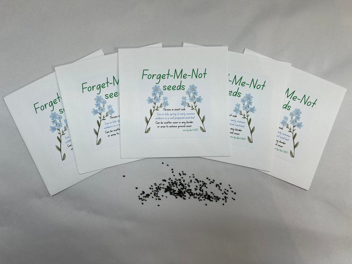 White envelopes are fanned out, each is titled “forget-me-not seeds”. There are pictures of blue forget-me-not flowers, and sowing instructions. There is a pile of small black seeds.