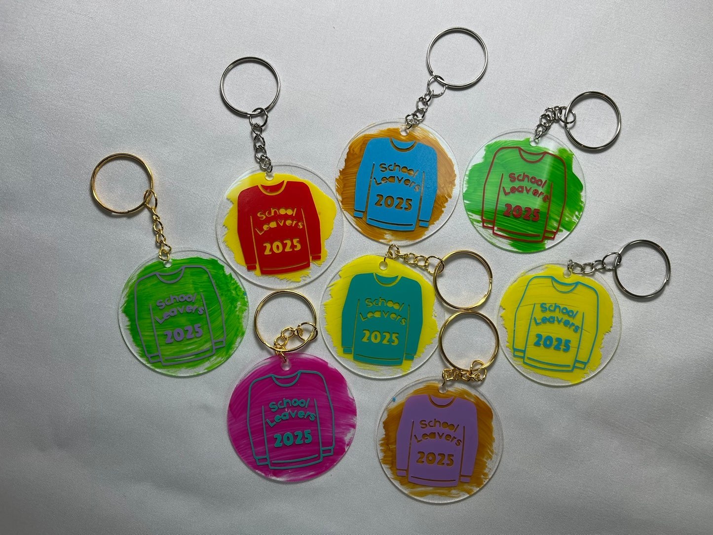 Circular acrylic keyrings are shown. They are painted in green, yellow, gold and pink. Each has a hoody on, printed with “school leavers 2025”. The printing is in red, blue, purple and aqua. The rings are a mixture of gold and silver. They look very colourful, each keyring is a unique mix of colours.