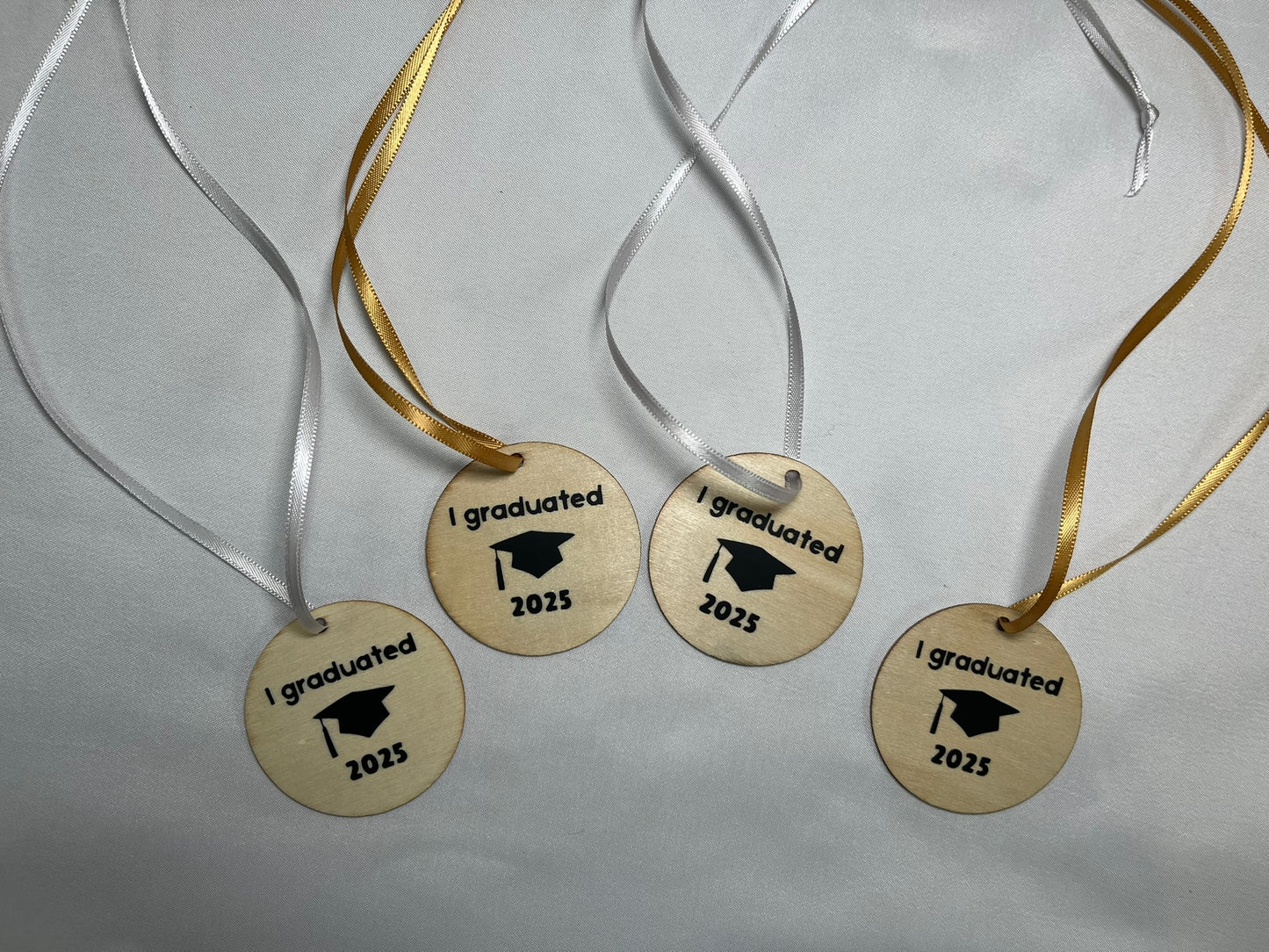 Four circular wooden medals sit on a white background. Some have silver ribbon, others have gold. The medals have a graduation cap on, and the words “I graduated 2025” on.