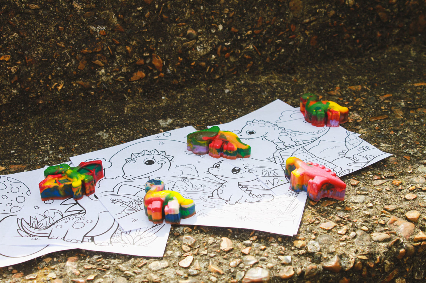 Five wax crayons in the shape of a dinosaurs sit on dinosaur colouring sheets. The crayons are green, yellow, orange, pink, blue and red. They are pterodactyl, diplodocus, t-rex and stegosaurus.