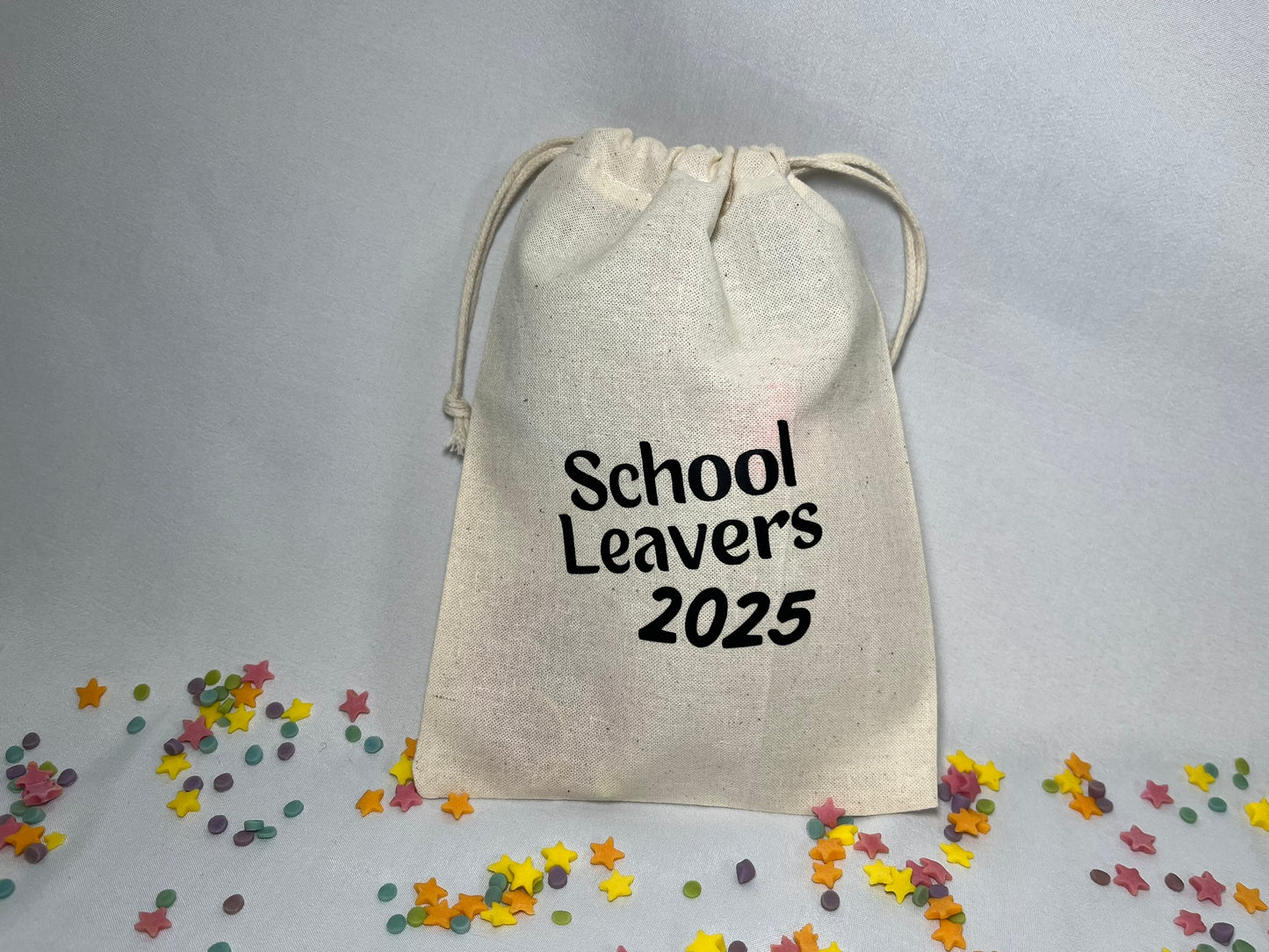A white canvas bag is on a bed of confetti and sprinkles. The bag is drawstring, and printed with the words “school leavers 2025”.