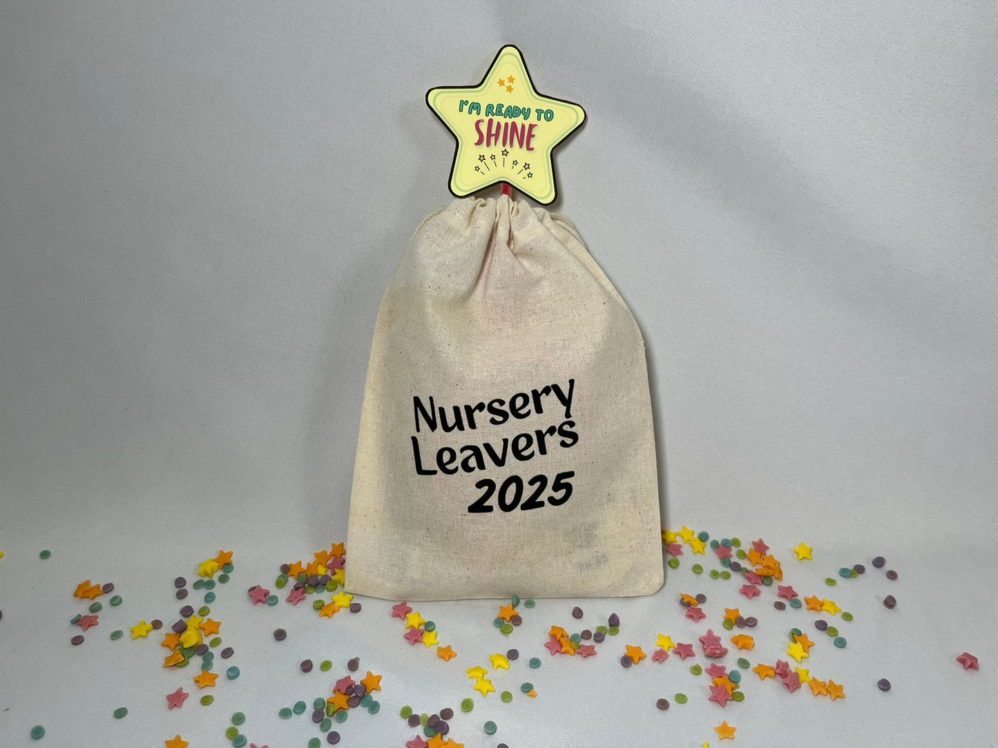 A white canvas bag is on a bed of confetti and sprinkles. The bag is drawstring, and printed with the words “nursery leavers 2025”. There is a star wand poking out the top.