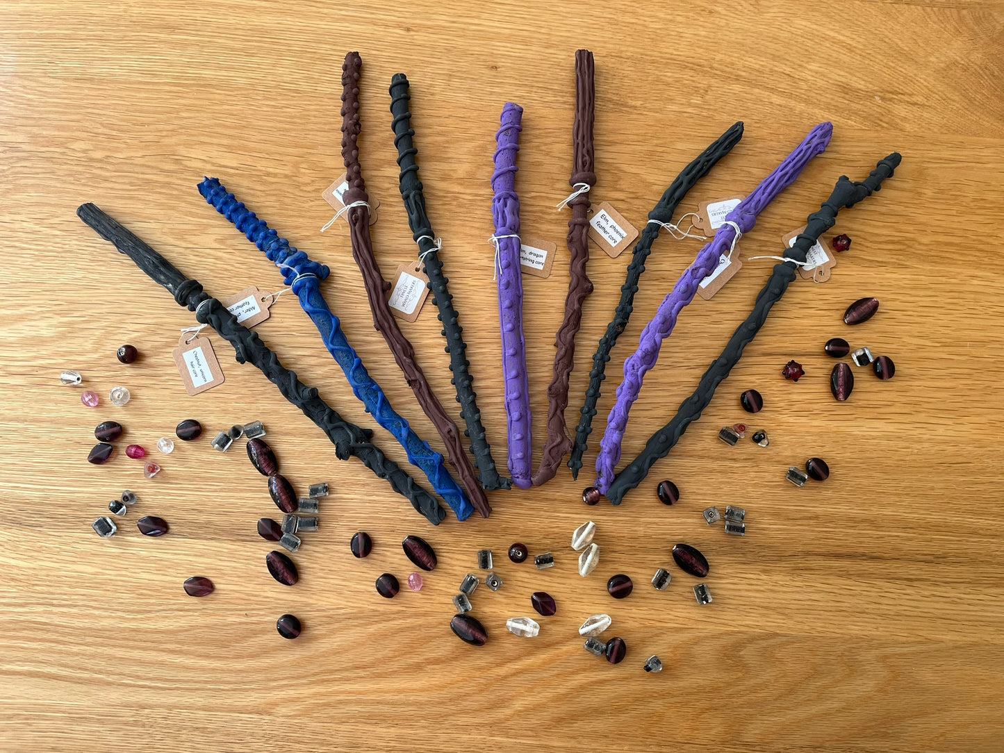 Nine wooden magic wands are fanned out on a wooden background. The wands are grey, blue, brown and purple, and have tags on saying "Finest Wand Makers". The wands are all different, and have different raised patterns. The wands are surrounded with shiny beads of different shapes and sizes.