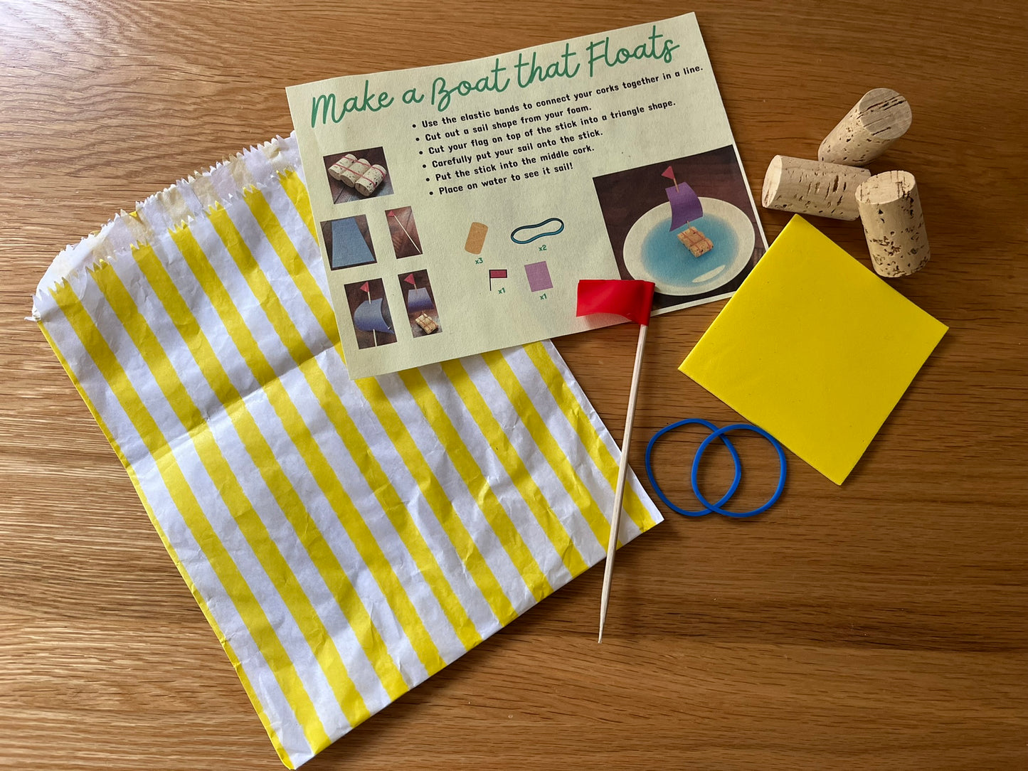 A square of yellow foam, blue elastic bands, three corks, a stick with a red flag at the end, a yellow striped paper bag and an instruction sheet for "Make a Boat that Floats". On a wooden background.
