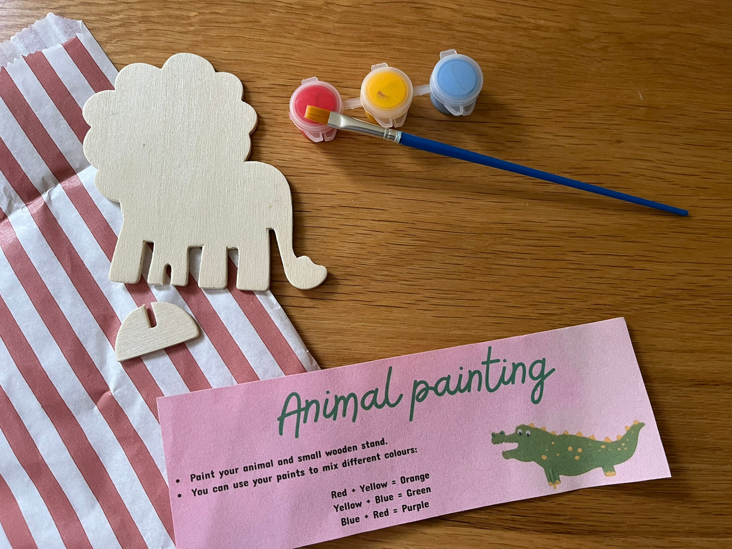 A red striped paper bag, instructions for "Animal Painting", a wooden lion shape, a stand for the lion, a paintbrush and small pots containing red, yellow and blue paint.