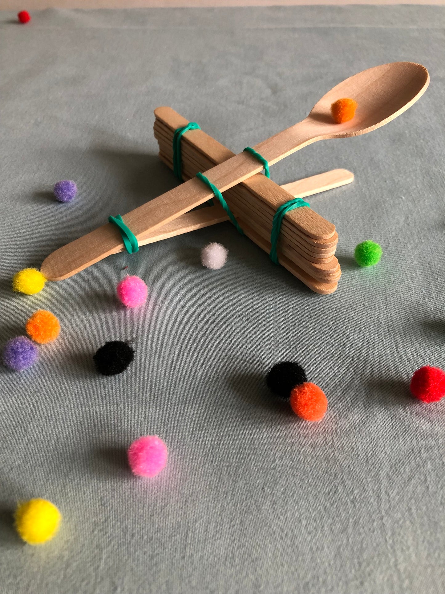 A wooden catapult made with a spoon lolly sticks and elastic bands is surrounded by rainbow pom poms.