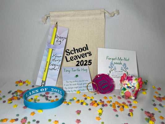A white canvas bag reading "School Leavers 2025" sits on a confetti background. The contents are shown: a yellow pencil printed with "dream big", a blue wristband that says Class of 2025, a turtle sits on a card with a poem titled Tiny Turtle Hug, a pink circular keyring has a school leavers 2025 hoody on it, there is a packet of Forget-Me-Not seeds, and a rainbow coloured axolotl squishy toy.