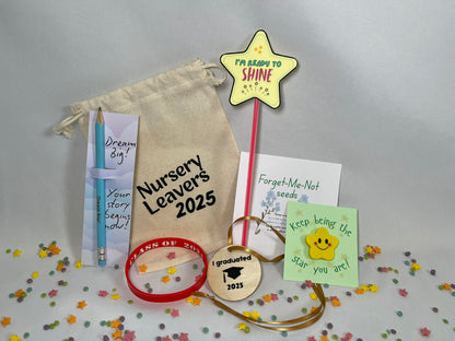 A white canvas bag reading "Nursery Leavers 2025" sits on a confetti background. The contents are shown: a blue pencil printed with "dream big", a red wristband that says Class of 2025, a yellow star rubber with a smile sits on a card titled “keep being the star you are”, a wooden circular medal has a graduation hat on it and the words “I graduated 2025”, there is a packet of Forget-Me-Not seeds. There’s a red glowstick with a star at the top, it looks like a magic wand, and reads Ready to Shine