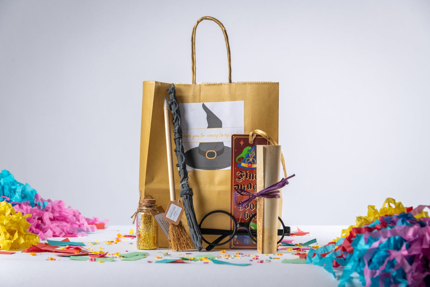 A grey wooden magic wand, a pencil that looks like a broomstick, a potion bottle with a cork top is filled with gold glitter, round Harry Potter wizard glasses, a scroll tied with purple raffia, a wizard bookmark. All sit in front of a gold paper party bag with a wizard witch hat sticker that reads “thank you for coming to my party”. They sit on a white background with rainbow confetti and sprinkles.