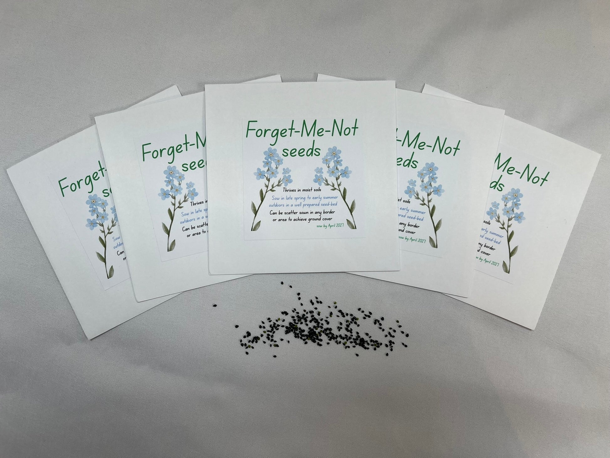 White envelopes are fanned out, each is titled “forget-me-not seeds”. There are pictures of blue forget-me-not flowers, and sowing instructions. There is a pile of small black seeds.