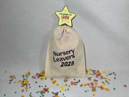 A white canvas bag is on a bed of confetti and sprinkles. The bag is drawstring, and printed with the words “nursery leavers 2025”. There is a star wand poking out the top.