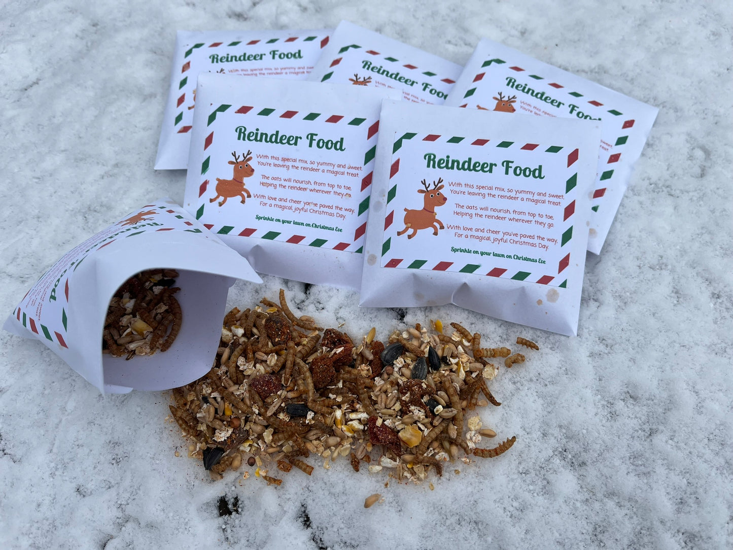 Five envelopes of Reindeer Food sit in the snow. In front there is an open packet with the contents spilled. You can see seeds, mealworms and cranberries.
