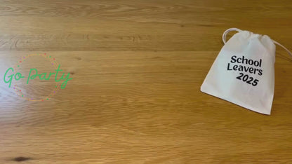 An unboxing video of the School Leavers 2025 bag: there is a blue class of 2025 wristband, a yellow pencil on a card saying dream big, a rainbow coloured axolotl squishy toy, a white envelope labelled forget-me-not seeds, a card with a turtle on titled tiny turtle hug, and a pink circular keyring that has a school leavers 2025 hoody on it. They are all laid out on a wooden surface.